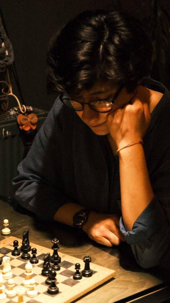 An adult concentrates deeply on a chess game in a dimly lit room, conveying strategy and focus.